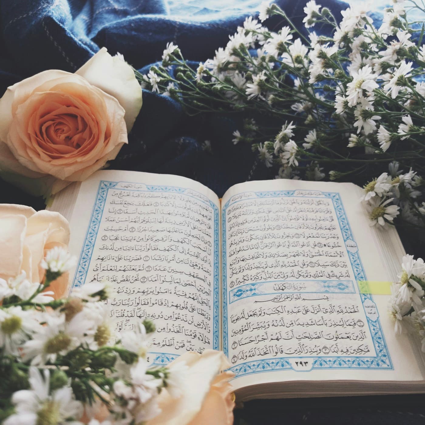 An open muṣḥaf at the start of Sūrat al-Kahf, resting among pale flowers.